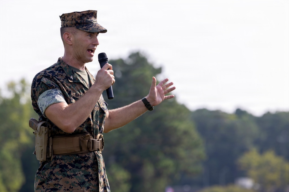 A U.S. Marine Corps officer in MARPAT digital camouflage uniform is speaking into a handheld microphone outdoors. He is gesturing with his right hand while addressing an unseen audience. The background shows blurred trees and an overcast sky, suggesting an outdoor military event or ceremony.