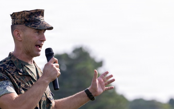 A U.S. Marine Corps officer in MARPAT digital camouflage uniform is speaking into a handheld microphone outdoors. He is gesturing with his right hand while addressing an unseen audience. The background shows blurred trees and an overcast sky, suggesting an outdoor military event or ceremony.