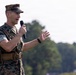 A U.S. Marine Corps officer in MARPAT digital camouflage uniform is speaking into a handheld microphone outdoors. He is gesturing with his right hand while addressing an unseen audience. The background shows blurred trees and an overcast sky, suggesting an outdoor military event or ceremony.