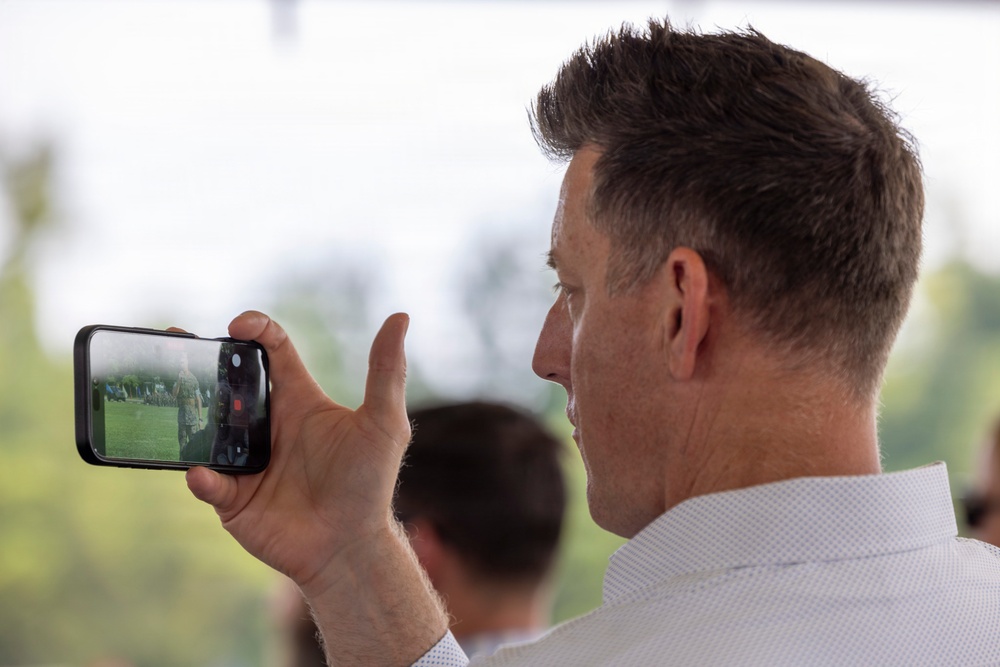 A middle-aged man with short brown hair is holding a smartphone horizontally to record or photograph an outdoor event. The phone screen displays what appears to be a person or figure on a green lawn. The image is taken from a rear three-quarter angle, with a blurred outdoor background.