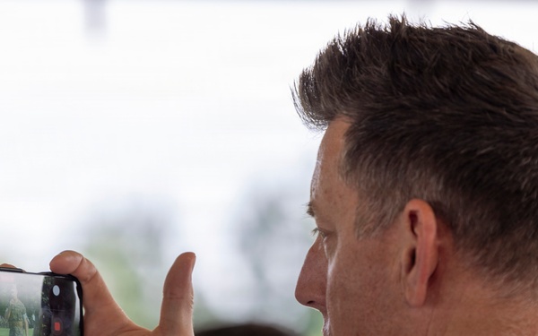 A middle-aged man with short brown hair is holding a smartphone horizontally to record or photograph an outdoor event. The phone screen displays what appears to be a person or figure on a green lawn. The image is taken from a rear three-quarter angle, with a blurred outdoor background.
