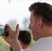 A middle-aged man with short brown hair is holding a smartphone horizontally to record or photograph an outdoor event. The phone screen displays what appears to be a person or figure on a green lawn. The image is taken from a rear three-quarter angle, with a blurred outdoor background.