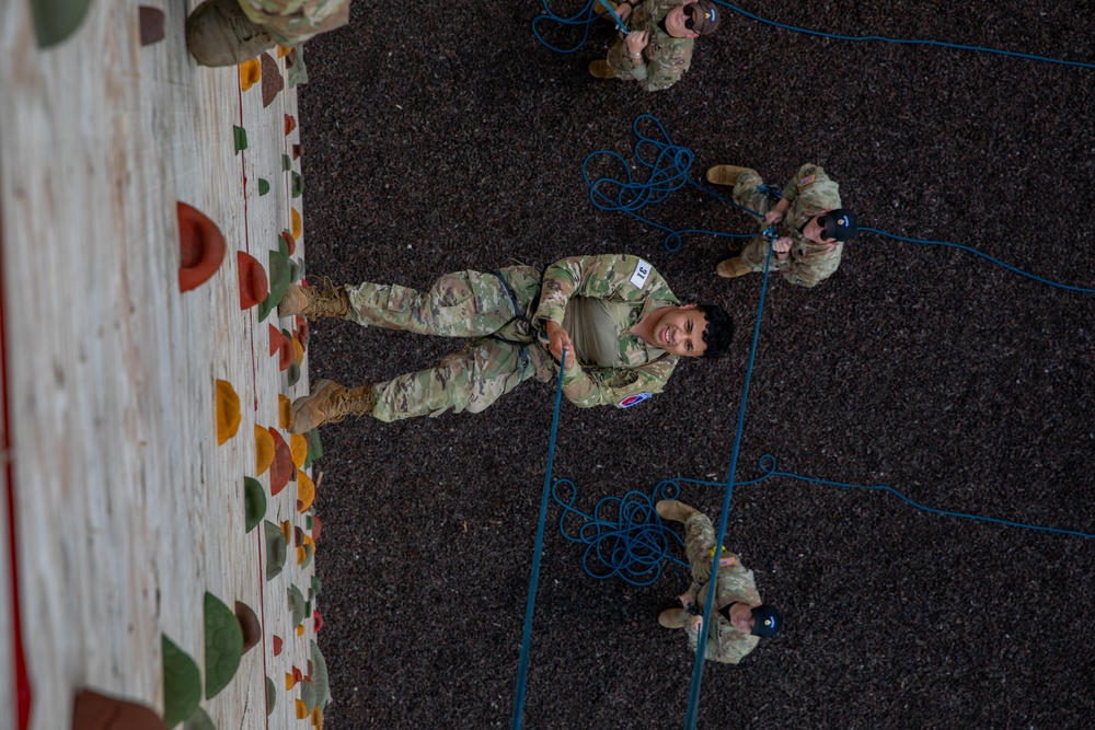 An overhead/top-down photograph showing military personnel in camouflage uniforms (OCP/MultiCam pattern) engaged in rappelling or rope training at what appears to be an indoor climbing wall facility. One central soldier is harnessed and connected to blue ropes while looking upward toward the camera, smiling. Several other soldiers on the ground are managing and belaying the ropes.