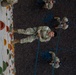 An overhead/top-down photograph showing military personnel in camouflage uniforms (OCP/MultiCam pattern) engaged in rappelling or rope training at what appears to be an indoor climbing wall facility. One central soldier is harnessed and connected to blue ropes while looking upward toward the camera, smiling. Several other soldiers on the ground are managing and belaying the ropes.