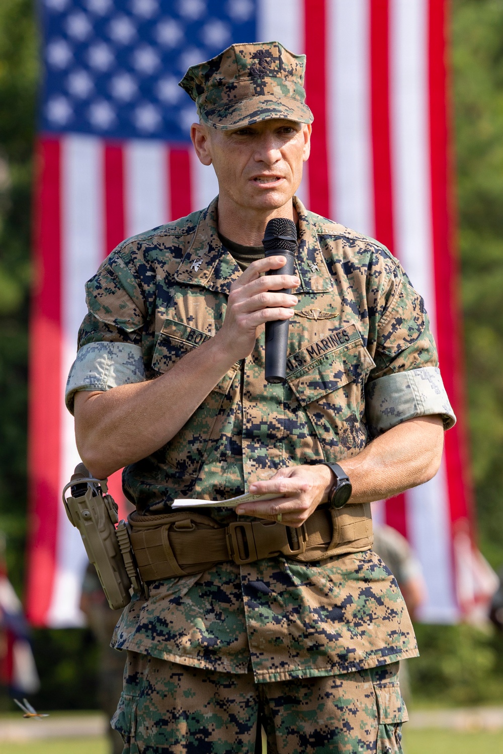 A U.S. Marine Corps officer in MARPAT digital camouflage uniform is speaking into a handheld microphone at what appears to be an official military ceremony or event. He is holding notes in his other hand and has a holstered sidearm on his left hip. A large American flag is prominently displayed in the background.