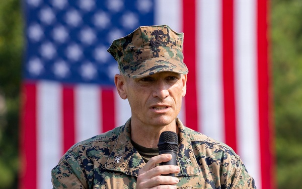 A U.S. Marine Corps officer in MARPAT digital camouflage uniform is speaking into a handheld microphone at what appears to be an official military ceremony or event. He is holding notes in his other hand and has a holstered sidearm on his left hip. A large American flag is prominently displayed in the background.