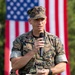 A U.S. Marine Corps officer in MARPAT digital camouflage uniform is speaking into a handheld microphone at what appears to be an official military ceremony or event. He is holding notes in his other hand and has a holstered sidearm on his left hip. A large American flag is prominently displayed in the background.