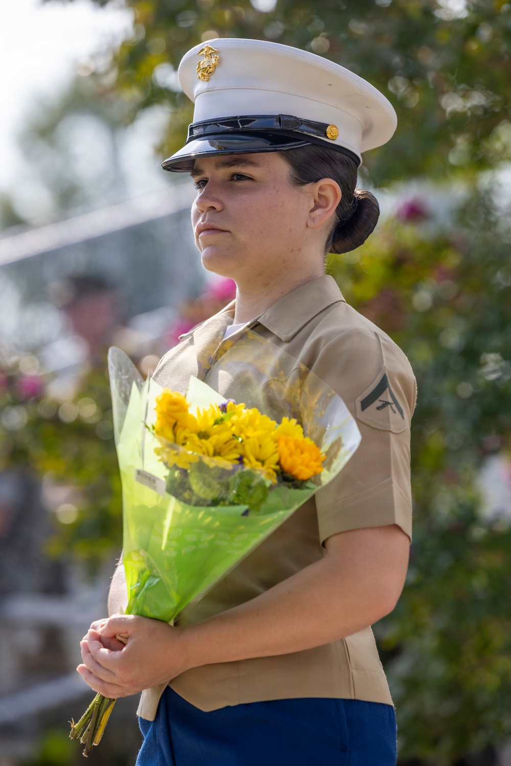 A female U.S. Marine in dress uniform stands at attention holding a bouquet of yellow and orange flowers wrapped in green cellophane. She wears the distinctive white cover (cap) with the Eagle, Globe, and Anchor emblem and a tan service uniform with a Private First Class chevron visible on her sleeve. Her expression is solemn and focused, suggesting a formal or ceremonial occasion.