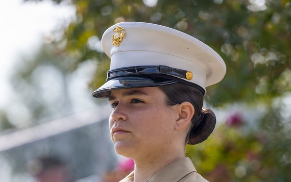 A female U.S. Marine in dress uniform stands at attention holding a bouquet of yellow and orange flowers wrapped in green cellophane. She wears the distinctive white cover (cap) with the Eagle, Globe, and Anchor emblem and a tan service uniform with a Private First Class chevron visible on her sleeve. Her expression is solemn and focused, suggesting a formal or ceremonial occasion.