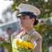 A female U.S. Marine in dress uniform stands at attention holding a bouquet of yellow and orange flowers wrapped in green cellophane. She wears the distinctive white cover (cap) with the Eagle, Globe, and Anchor emblem and a tan service uniform with a Private First Class chevron visible on her sleeve. Her expression is solemn and focused, suggesting a formal or ceremonial occasion.