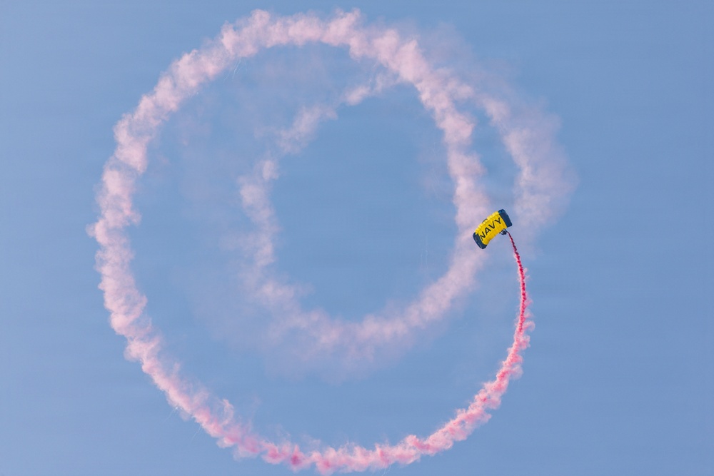 A Navy parachutist with a yellow canopy labeled 'NAVY' is performing an aerial demonstration, trailing red/pink colored smoke that has formed a near-complete circular loop in a clear blue sky. The smoke trail indicates the jumper has completed approximately one full circular maneuver. This appears to be a military airshow or public demonstration performance.