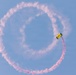 A Navy parachutist with a yellow canopy labeled 'NAVY' is performing an aerial demonstration, trailing red/pink colored smoke that has formed a near-complete circular loop in a clear blue sky. The smoke trail indicates the jumper has completed approximately one full circular maneuver. This appears to be a military airshow or public demonstration performance.