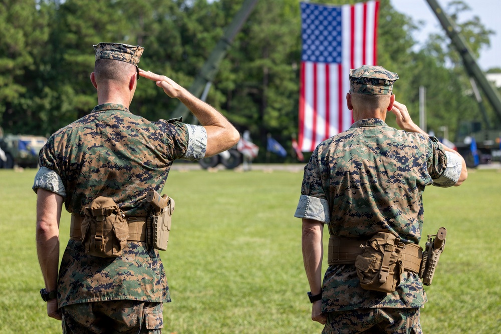 Two U.S. Marine Corps personnel in MARPAT woodland camouflage uniforms are photographed from behind as they render a salute toward a large American flag. The setting appears to be an outdoor military ceremony on a grass parade field. Both individuals are in full utility uniform with tactical gear attached to their belts.