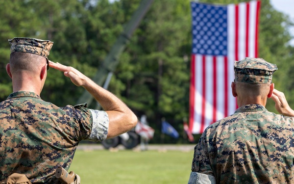 Two U.S. Marine Corps personnel in MARPAT woodland camouflage uniforms are photographed from behind as they render a salute toward a large American flag. The setting appears to be an outdoor military ceremony on a grass parade field. Both individuals are in full utility uniform with tactical gear attached to their belts.