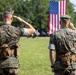 Two U.S. Marine Corps personnel in MARPAT woodland camouflage uniforms are photographed from behind as they render a salute toward a large American flag. The setting appears to be an outdoor military ceremony on a grass parade field. Both individuals are in full utility uniform with tactical gear attached to their belts.