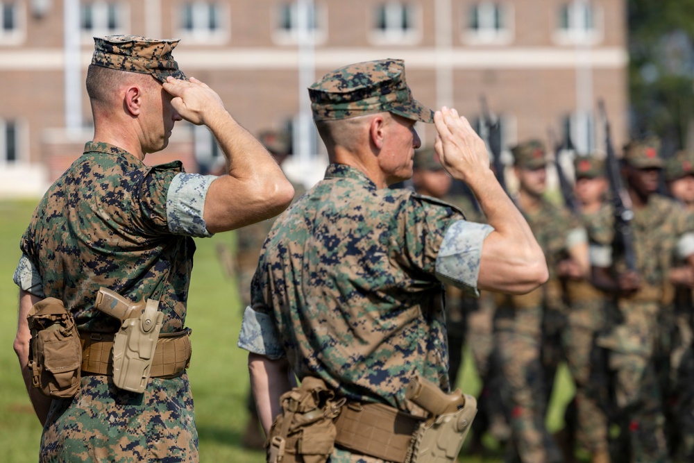 Two U.S. Marine Corps officers in the foreground are rendering a formal salute during what appears to be a military ceremony or parade. Both are wearing MARPAT (Marine Pattern) digital camouflage uniforms with covers (caps). In the background, a formation of Marines in similar uniforms can be seen holding rifles at attention.
