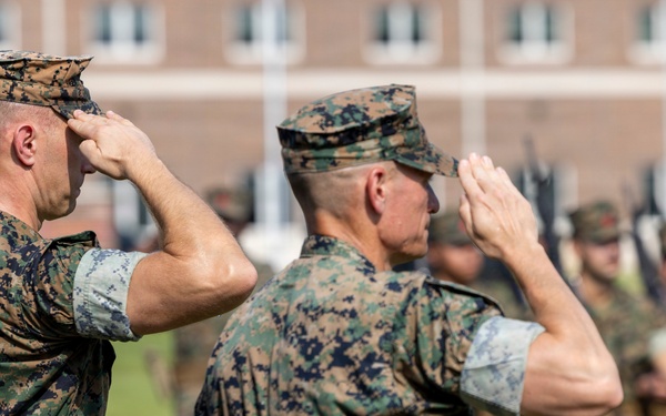 Two U.S. Marine Corps officers in the foreground are rendering a formal salute during what appears to be a military ceremony or parade. Both are wearing MARPAT (Marine Pattern) digital camouflage uniforms with covers (caps). In the background, a formation of Marines in similar uniforms can be seen holding rifles at attention.