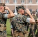 Two U.S. Marine Corps officers in the foreground are rendering a formal salute during what appears to be a military ceremony or parade. Both are wearing MARPAT (Marine Pattern) digital camouflage uniforms with covers (caps). In the background, a formation of Marines in similar uniforms can be seen holding rifles at attention.