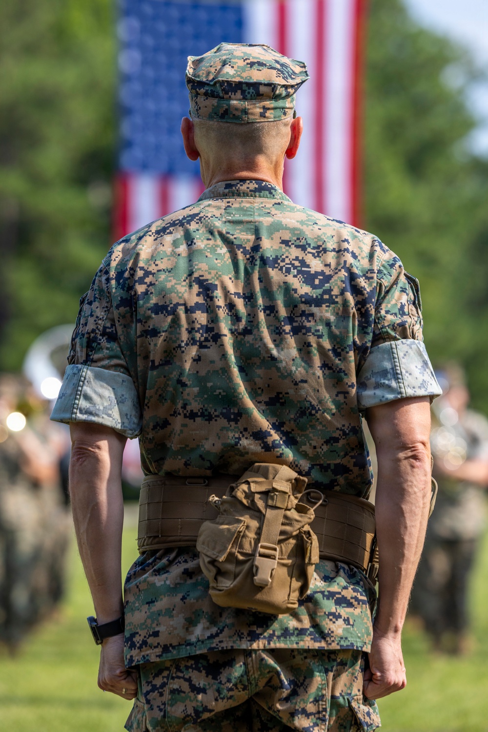 A U.S. Marine in MARPAT digital camouflage uniform stands at attention facing away from the camera, with a large American flag displayed prominently in the background. The setting appears to be an outdoor military ceremony or parade, with what appears to be a military band visible in the blurred background. The composition emphasizes patriotism and military formality.