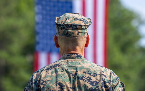 A U.S. Marine in MARPAT digital camouflage uniform stands at attention facing away from the camera, with a large American flag displayed prominently in the background. The setting appears to be an outdoor military ceremony or parade, with what appears to be a military band visible in the blurred background. The composition emphasizes patriotism and military formality.