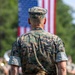 A U.S. Marine in MARPAT digital camouflage uniform stands at attention facing away from the camera, with a large American flag displayed prominently in the background. The setting appears to be an outdoor military ceremony or parade, with what appears to be a military band visible in the blurred background. The composition emphasizes patriotism and military formality.