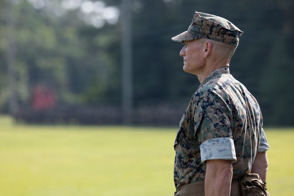 A male military service member in U.S. Marine Corps MARPAT (Marine Pattern) digital camouflage uniform is photographed in profile view. He is standing at attention or in an observational stance on what appears to be a grassy field or parade ground. The image has a shallow depth of field with a blurred green background of trees and grass.