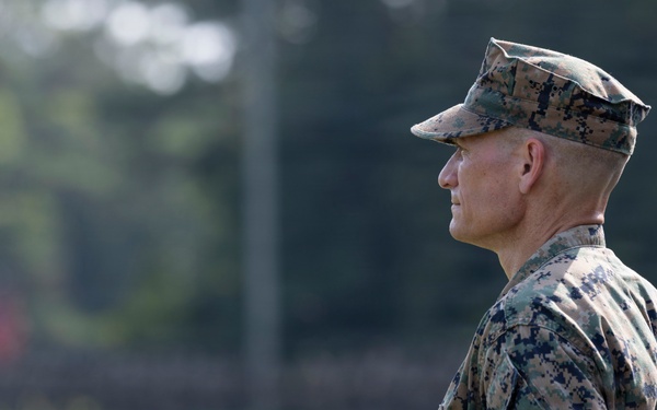 A male military service member in U.S. Marine Corps MARPAT (Marine Pattern) digital camouflage uniform is photographed in profile view. He is standing at attention or in an observational stance on what appears to be a grassy field or parade ground. The image has a shallow depth of field with a blurred green background of trees and grass.