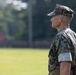 A male military service member in U.S. Marine Corps MARPAT (Marine Pattern) digital camouflage uniform is photographed in profile view. He is standing at attention or in an observational stance on what appears to be a grassy field or parade ground. The image has a shallow depth of field with a blurred green background of trees and grass.