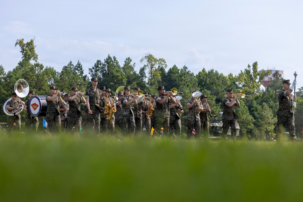 A U.S. Marine Corps marching band performs outdoors on a grassy field, wearing woodland MARPAT camouflage uniforms. The band includes brass, woodwind, and percussion instruments, with a drum major leading at the right side. A bass drum bearing what appears to be a Marine Corps divisional insignia is visible on the left.