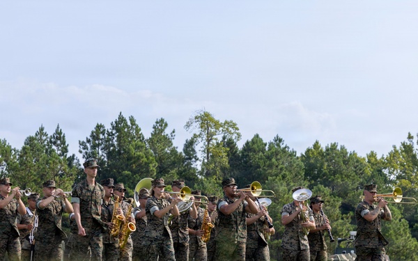 A U.S. Marine Corps marching band performs outdoors on a grassy field, wearing woodland MARPAT camouflage uniforms. The band includes brass, woodwind, and percussion instruments, with a drum major leading at the right side. A bass drum bearing what appears to be a Marine Corps divisional insignia is visible on the left.