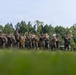 A U.S. Marine Corps marching band performs outdoors on a grassy field, wearing woodland MARPAT camouflage uniforms. The band includes brass, woodwind, and percussion instruments, with a drum major leading at the right side. A bass drum bearing what appears to be a Marine Corps divisional insignia is visible on the left.