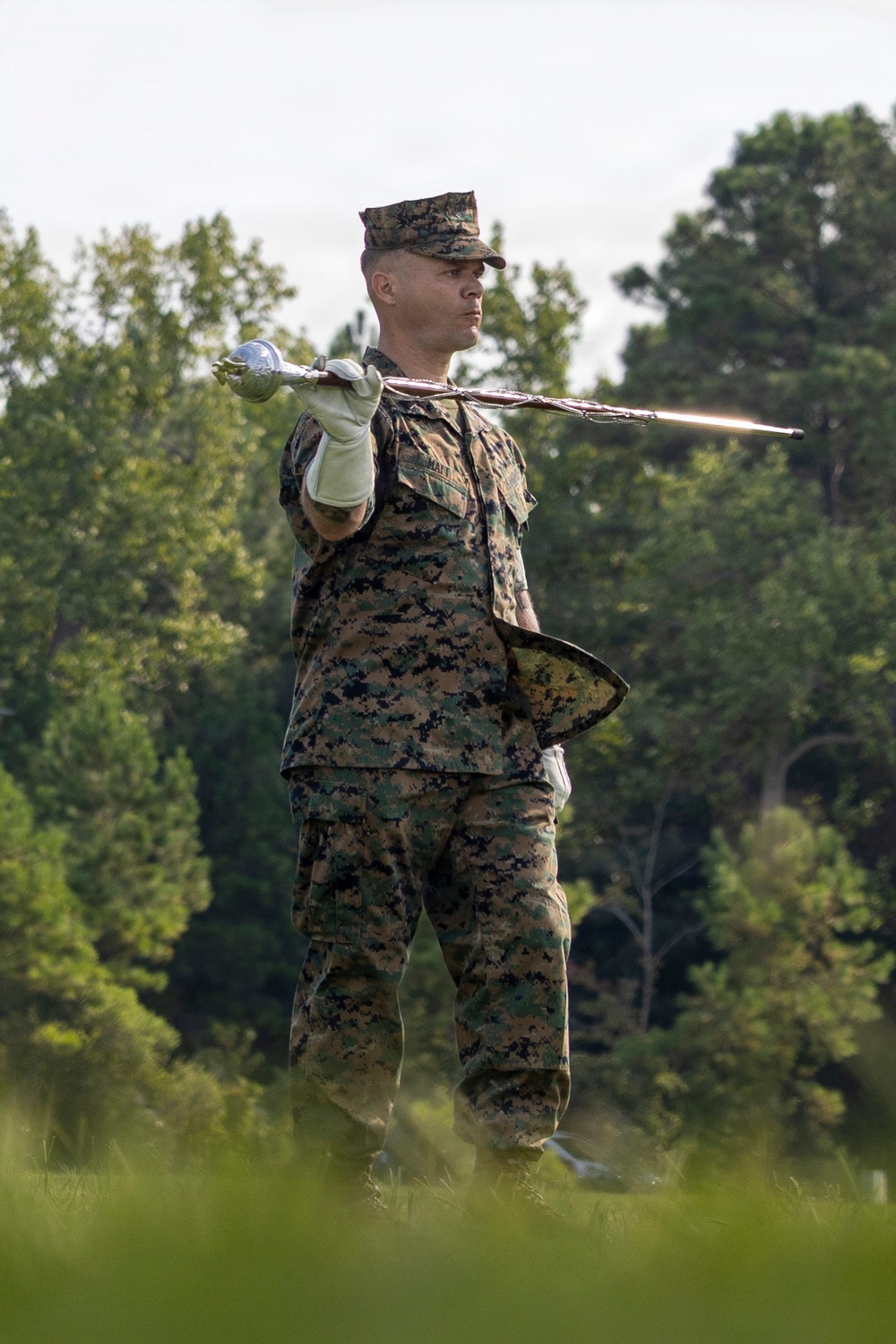 A U.S. Marine in MARPAT woodland camouflage uniform is holding a ceremonial sword extended outward at shoulder height with a white-gloved hand, while holding what appears to be a ceremonial guidon or flag base in the other hand. The Marine is standing in an outdoor setting with lush green trees in the background, suggesting a formal military ceremony or drill practice. The posture and equipment indicate a color guard or ceremonial drill team activity.