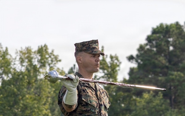 A U.S. Marine in MARPAT woodland camouflage uniform is holding a ceremonial sword extended outward at shoulder height with a white-gloved hand, while holding what appears to be a ceremonial guidon or flag base in the other hand. The Marine is standing in an outdoor setting with lush green trees in the background, suggesting a formal military ceremony or drill practice. The posture and equipment indicate a color guard or ceremonial drill team activity.