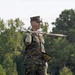 A U.S. Marine in MARPAT woodland camouflage uniform is holding a ceremonial sword extended outward at shoulder height with a white-gloved hand, while holding what appears to be a ceremonial guidon or flag base in the other hand. The Marine is standing in an outdoor setting with lush green trees in the background, suggesting a formal military ceremony or drill practice. The posture and equipment indicate a color guard or ceremonial drill team activity.