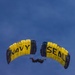 Two Navy SEAL parachutists are performing a formation skydive against a clear blue sky. Their parachutes are yellow and black, prominently displaying 'NAVY' and 'SEAL' text respectively. The two canopies appear to be flying in close proximity, suggesting a coordinated aerial demonstration.