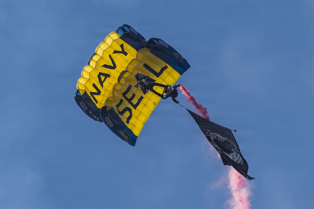 A Navy SEAL parachutist descends under a large yellow and navy blue parachute emblazoned with 'NAVY SEAL' text. The jumper is trailing red smoke and carrying a black flag, likely during an airshow or ceremonial demonstration. The image is captured from a low angle against a clear blue sky.