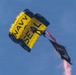 A Navy SEAL parachutist descends under a large yellow and navy blue parachute emblazoned with 'NAVY SEAL' text. The jumper is trailing red smoke and carrying a black flag, likely during an airshow or ceremonial demonstration. The image is captured from a low angle against a clear blue sky.