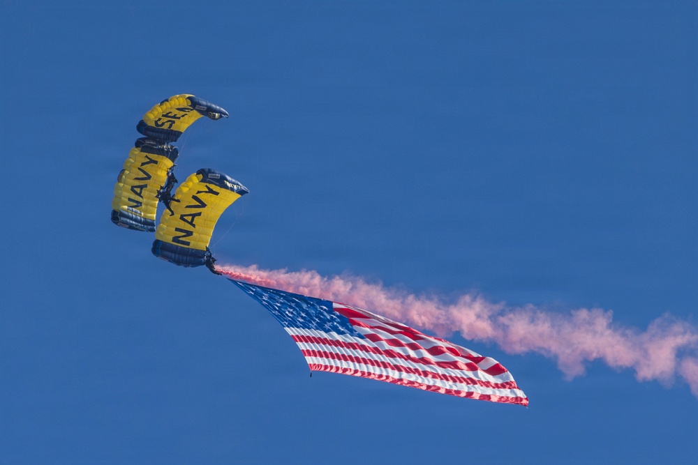 Two U.S. Navy parachutists are descending under yellow and navy blue canopies marked with 'NAVY' and 'SEAL' text against a clear blue sky. They are carrying a large American flag while emitting pink/red colored smoke. This appears to be a demonstration jump, likely at an airshow or military ceremony.