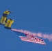 Two U.S. Navy parachutists are descending under yellow and navy blue canopies marked with 'NAVY' and 'SEAL' text against a clear blue sky. They are carrying a large American flag while emitting pink/red colored smoke. This appears to be a demonstration jump, likely at an airshow or military ceremony.