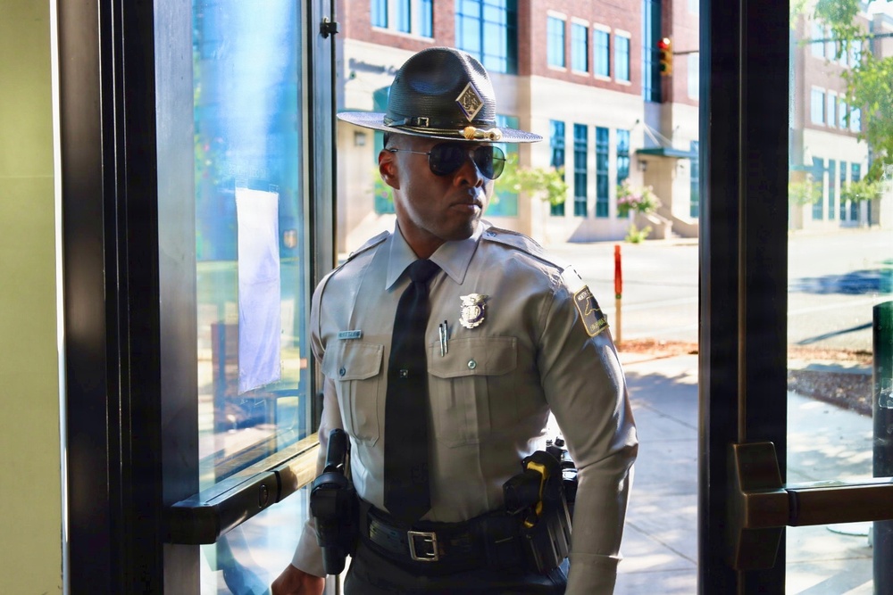 A law enforcement officer in a light gray uniform stands at a glass entrance doorway, appearing to be on duty. He is wearing a campaign/drill sergeant-style hat, aviator sunglasses, a dark tie, and a badge on his chest. The background shows an urban street scene with brick buildings and a traffic light.