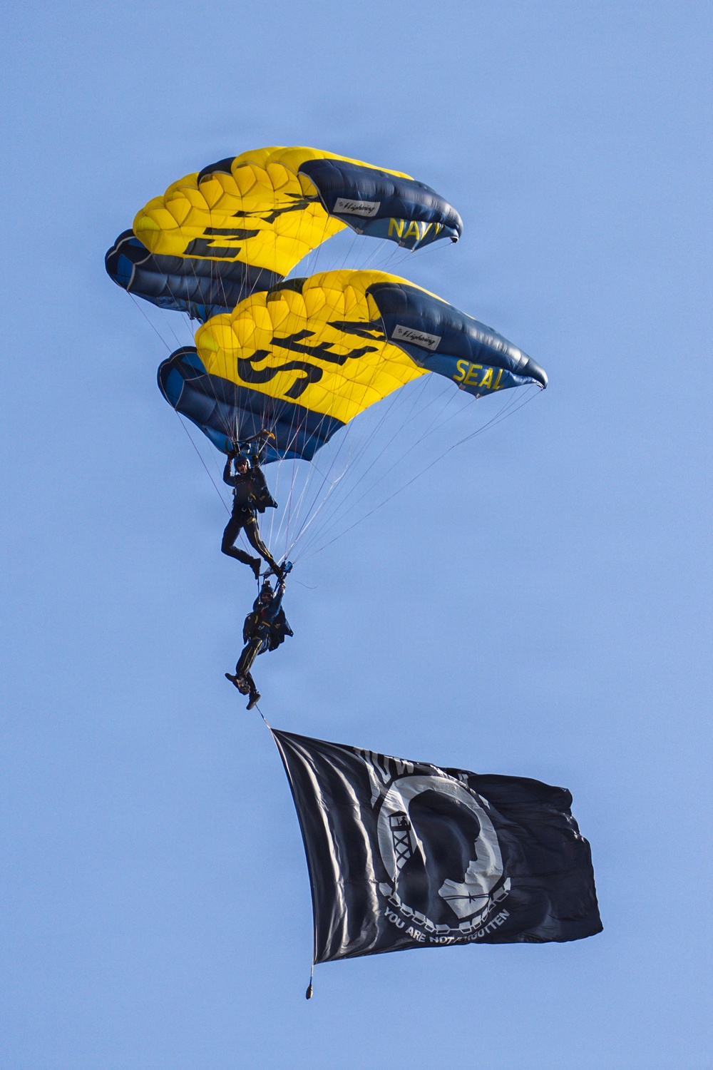 Two skydivers from the U.S. Navy SEAL parachute demonstration team descend under stacked yellow and navy blue canopies marked with 'NAVY' and 'SEAL' branding. The lower jumper is carrying a large black POW/MIA flag reading 'YOU ARE NOT FORGOTTEN,' trailing below them against a clear blue sky. This appears to be a ceremonial military demonstration jump.