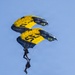 Two skydivers from the U.S. Navy SEAL parachute demonstration team descend under stacked yellow and navy blue canopies marked with 'NAVY' and 'SEAL' branding. The lower jumper is carrying a large black POW/MIA flag reading 'YOU ARE NOT FORGOTTEN,' trailing below them against a clear blue sky. This appears to be a ceremonial military demonstration jump.