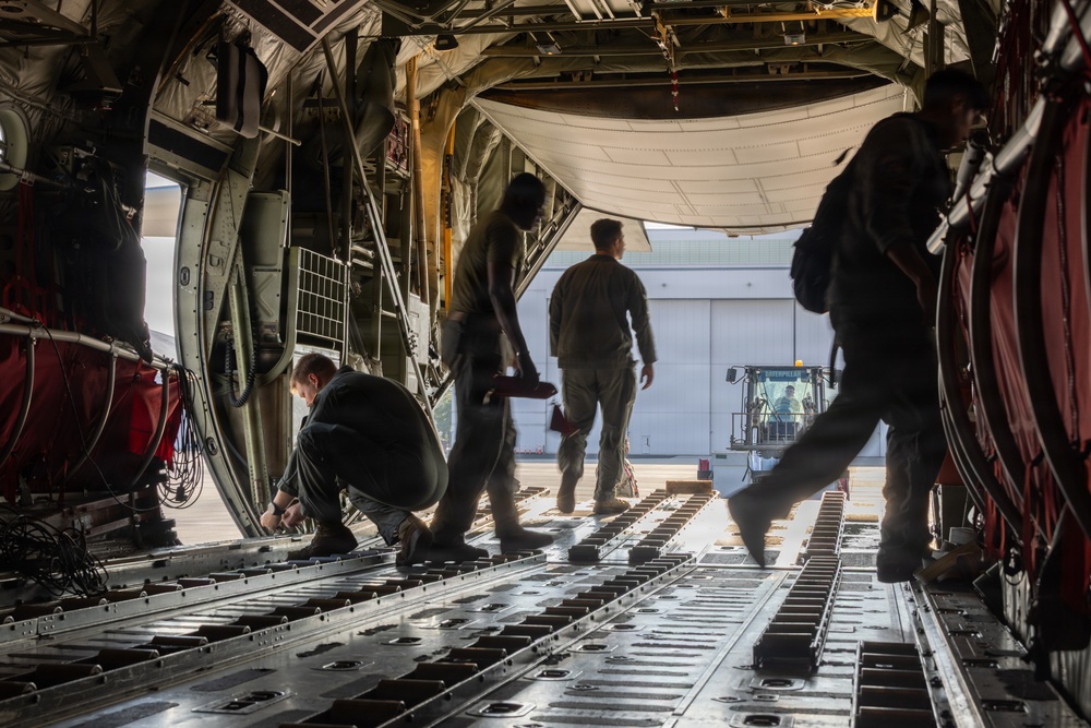 Military personnel are working inside the cargo hold of a large military transport aircraft, likely a C-130 Hercules or similar airlifter. The crew members appear to be preparing the aircraft for loading or unloading operations, with one person crouching to work on the cargo floor while others stand near the open rear ramp. A ground support vehicle, identifiable as a Caterpillar machine, is visible outside on the tarmac.