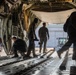 Military personnel are working inside the cargo hold of a large military transport aircraft, likely a C-130 Hercules or similar airlifter. The crew members appear to be preparing the aircraft for loading or unloading operations, with one person crouching to work on the cargo floor while others stand near the open rear ramp. A ground support vehicle, identifiable as a Caterpillar machine, is visible outside on the tarmac.