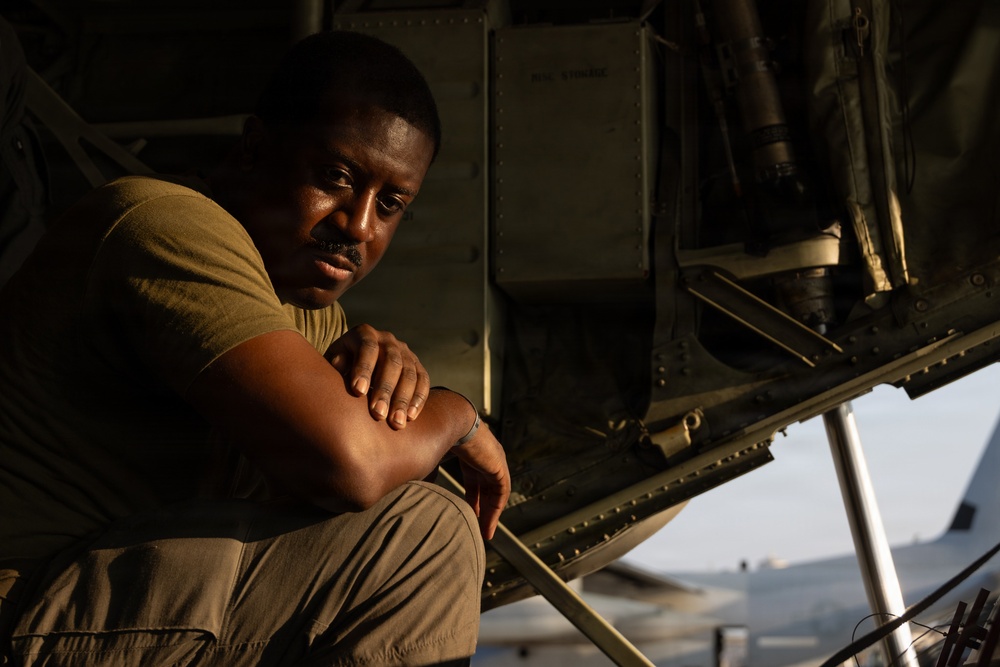 A man in military-style olive drab clothing sits beneath what appears to be a large military aircraft, resting with his arms crossed over his knees. He is looking directly at the camera with a composed, contemplative expression. The image is lit with warm, directional light suggesting early morning or late afternoon conditions.