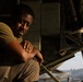 A man in military-style olive drab clothing sits beneath what appears to be a large military aircraft, resting with his arms crossed over his knees. He is looking directly at the camera with a composed, contemplative expression. The image is lit with warm, directional light suggesting early morning or late afternoon conditions.