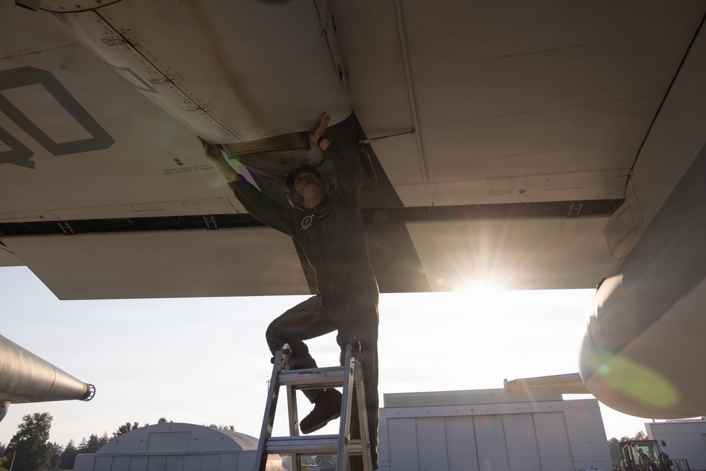 A technician or maintenance crew member in an olive/green flight suit is working on the underside of a large aircraft wing while standing on a metal stepladder. The photo is taken from a low angle looking upward, with strong sunlight creating a lens flare effect in the background. The setting appears to be an outdoor military or aviation maintenance area.