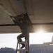 A technician or maintenance crew member in an olive/green flight suit is working on the underside of a large aircraft wing while standing on a metal stepladder. The photo is taken from a low angle looking upward, with strong sunlight creating a lens flare effect in the background. The setting appears to be an outdoor military or aviation maintenance area.