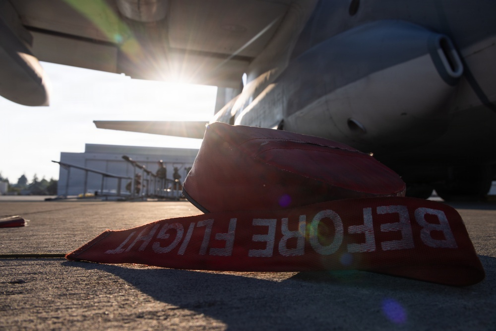 A ground-level photograph taken beneath a large military or transport aircraft on a tarmac. Red wheel chocks with 'BEFORE FLIGHT' text are visible in the foreground, indicating pre-flight ground safety equipment. The image is backlit by a bright sun flare, with personnel and a hangar structure visible in the background.