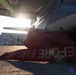 A ground-level photograph taken beneath a large military or transport aircraft on a tarmac. Red wheel chocks with 'BEFORE FLIGHT' text are visible in the foreground, indicating pre-flight ground safety equipment. The image is backlit by a bright sun flare, with personnel and a hangar structure visible in the background.